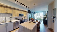 This stylish single-floor urban residence combines contemporary design with functional living. The open-concept kitchen serves as the heart of the space, outfitted with stainless steel appliances including a French door refrigerator, gas range, built-in microwave, and dishwasher. Light wood cabinetry with sleek hardware sits above a white subway tile backsplash, while lower grey cabinets and white quartz countertops complete the refined palette. A spacious island with bar seating and pendant lighting makes the kitchen both practical and inviting. Track lighting runs the length of the kitchen ceiling, adding focused illumination throughout.

The living area flows naturally from the kitchen and is anchored by floor-to-ceiling sliding glass doors that open to a private balcony, offering expansive city skyline views and an abundance of natural light. Light wood-tone flooring runs throughout, lending warmth and continuity to the open layout.

The bedroom is a serene retreat, featuring a large window with urban views, warm-toned hardwood floors, and soft neutral walls. An adjacent walk-in closet provides generous open shelving and hanging storage. The bathroom is finished with white subway tile surrounding a soaking tub and shower, a wide vanity with quartz countertop, gold fixtures, and a large framed mirror.