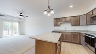 This single-floor residence offers a thoughtfully designed layout that balances functionality and comfort throughout its living spaces. The kitchen serves as a central highlight, showcasing rich dark-toned cabinetry, granite countertops, and a full suite of stainless steel appliances including a refrigerator, electric range with overhead microwave, and dishwasher. A generously sized island with matching granite provides additional prep space and casual seating, illuminated by an elegant pendant chandelier overhead. Light hardwood-style flooring runs throughout the kitchen and extends into adjoining areas.

The living area features plush neutral carpeting and a ceiling fan, with access to an outdoor balcony framed by views of mature trees, bringing natural light and greenery into the space. Bedrooms are carpeted and bright, with large windows offering outdoor views and ample closet space accessed via sliding doors. The bathroom includes a full soaking tub and shower combination with clean white surrounds and light wood-style flooring. A dedicated laundry room with full-size washer and dryer units and wire shelving overhead adds everyday convenience. Overall, the space presents a clean, modern aesthetic with neutral tones throughout.
