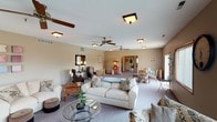 This single-floor community space is thoughtfully designed to accommodate a variety of social activities and gatherings. The open floor plan is anchored by a generous main lounge area featuring plush sofas, accent chairs, and glass-topped coffee tables arranged to encourage conversation and comfort. Multiple ceiling fans throughout ensure consistent airflow and comfort across the expansive layout.

A standout feature of the space is a cozy sitting room with a fireplace set against a rich, warm-toned accent wall, accompanied by a wall-mounted television — an inviting retreat for relaxed viewing and socializing. Soft, neutral carpeting flows throughout, complementing the warm beige tones of the walls and the wood-trimmed finishes.

Additional seating nooks offer quieter settings, furnished with upholstered armchairs, console tables, and decorative mirrors that add depth and warmth to the space. A charming popcorn machine adds a fun, communal touch, hinting at the space's role as a social hub. Large windows along one wall welcome natural light and provide pleasant outward views, enhancing the open and airy atmosphere. The textured ceilings and flush-mount lighting fixtures complete the well-maintained, welcoming character of this versatile community gathering space.