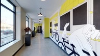This single-floor space makes a memorable first impression through its expansive hallway, which serves as both a functional passage and an artistic showcase. The corridor is dominated by a large-scale illustrated mural depicting whimsical characters — including dogs riding bicycles — rendered in bold black, white, and yellow tones against a bright chartreuse background. The mural spans the full length of one wall, creating a dynamic and visually engaging environment.

The hallway features polished concrete flooring that reflects light and contributes to the space's modern, urban aesthetic. A row of industrial-style pendant lights hangs from the ceiling at regular intervals, providing consistent illumination throughout the corridor while reinforcing the contemporary design language. Natural light filters in through windows along the opposite wall, complementing the artificial lighting.

Large, dark rectangular planters filled with lush foliage are positioned along the windowed wall, introducing an organic element that balances the graphic intensity of the mural. The combination of artistic expression, thoughtful lighting, greenery, and clean architectural lines gives this space a creative, welcoming, and design-forward character.