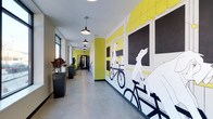 This single-floor space makes a memorable first impression through its expansive hallway, which serves as both a functional passage and an artistic showcase. The corridor is dominated by a large-scale illustrated mural depicting whimsical characters — including dogs riding bicycles — rendered in bold black, white, and yellow tones against a bright chartreuse background. The mural spans the full length of one wall, creating a dynamic and visually engaging environment.

The hallway features polished concrete flooring that reflects light and contributes to the space's modern, urban aesthetic. A row of industrial-style pendant lights hangs from the ceiling at regular intervals, providing consistent illumination throughout the corridor while reinforcing the contemporary design language. Natural light filters in through windows along the opposite wall, complementing the artificial lighting.

Large, dark rectangular planters filled with lush foliage are positioned along the windowed wall, introducing an organic element that balances the graphic intensity of the mural. The combination of artistic expression, thoughtful lighting, greenery, and clean architectural lines gives this space a creative, welcoming, and design-forward character.