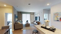 This single-floor residence showcases a well-designed open-concept layout that seamlessly connects the kitchen and living areas. The living room features warm hardwood-style flooring, neutral gray walls, and a large sliding glass door fitted with vertical blinds that floods the space with natural light. The living area comfortably accommodates a sectional sofa, ottoman, and entertainment center, creating a welcoming and functional gathering space. Decorative wall art adds character to the room, while the layout provides clear sightlines into an adjacent bedroom. The kitchen area, partially visible in the foreground, highlights elegant granite countertops with attractive veining and a breakfast bar with seating. Ceiling-mounted HVAC vents are integrated throughout, ensuring climate comfort. The overall color palette of soft grays, warm wood tones, and teal accents creates a cohesive, modern aesthetic. The open flow between rooms enhances the sense of spaciousness, making this a practical and visually appealing single-level living space.