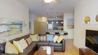 This single-floor residence offers an open-concept design that seamlessly connects the living area and kitchen, creating a cohesive and functional layout. The living room is anchored by a large dark brown sectional sofa adorned with colorful accent pillows in teal, green, and multicolored patterns, complemented by a wood coffee table. A ceiling fan with a light fixture overhead provides both airflow and ambient lighting. Warm wood-look flooring runs throughout the space, adding a welcoming feel.

The kitchen is positioned at the rear of the open layout and features dark cabinetry, a stainless steel refrigerator, and a kitchen island or breakfast bar with bar-height seating. Three pendant lights hang above the bar area, providing focused task lighting, while recessed lighting illuminates the kitchen workspace. The dark cabinetry contrasts with lighter countertops, giving the kitchen a modern aesthetic.

A hallway visible to the left leads to additional rooms, with louvered closet doors and an entry door adding architectural detail. Wall art and decorative accents contribute to the overall inviting and contemporary ambiance of the space.
