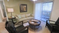 This single-floor residential unit offers a comfortable and thoughtfully designed living environment. The living room serves as the heart of the space, featuring warm, neutral-toned walls that create a calm and inviting atmosphere. Plush carpeted flooring runs throughout, adding to the overall sense of comfort and warmth. A generously sized sofa paired with accent chairs provides ample seating, centered around a distinctive round wicker coffee table that adds a natural, earthy element to the room's décor. Natural light flows into the space through a large sliding glass door dressed with vertical blinds, which opens to an outdoor area — a standout feature that extends the living space beyond the interior. Overhead, a contemporary chandelier-style light fixture adds a refined touch to the room. The walls are accented with framed artwork, contributing to the polished, move-in-ready aesthetic. The overall ambiance of the unit is warm, cozy, and well-lit, with a design sensibility that balances casual comfort with tasteful styling, making it a functional and visually appealing living space.