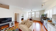 This single-floor residence offers a thoughtfully designed layout with a clean, contemporary aesthetic throughout. Rich hardwood floors in a warm reddish-brown tone run continuously through the main living areas, adding warmth and character to the space.

The open-concept living room is generously sized, featuring tall ceilings, recessed lighting, and a ceiling fan. A cozy seating arrangement centers around a fireplace media console, creating an inviting atmosphere for relaxing or entertaining.

Adjacent to the living area, the dining space benefits from an abundance of natural light streaming through multiple large windows that frame views of the surrounding neighborhood. The bright, airy feel carries into the well-equipped kitchen, which features white shaker cabinetry, granite-style countertops, and a full suite of stainless steel appliances including a refrigerator, electric range, and built-in microwave.

The bedroom is a calm, comfortable retreat with dual windows offering ample natural light and neighborhood views, complemented by neutral-toned walls that create a relaxing environment. The full bathroom is finished with matching granite-style countertops, white cabinetry, and a shower/tub combination. Overall, the space presents a cohesive, light-filled interior with practical finishes throughout.