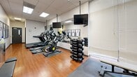 Located at 4830 Cass Ave in Detroit, Michigan, this fitness center offers a thoughtfully organized workout space within a single open room. The layout features warm hardwood flooring that transitions to rubber matting in the strength training zone, providing both comfort and functionality underfoot. A drop ceiling with recessed panel lighting ensures consistent, even illumination throughout.

The cardio section is lined with Star Trac treadmills, elliptical trainers, and a stationary cycling bike, each equipped with mounted flat-screen TVs for entertainment during workouts. A multi-station cable machine anchors the strength training area, complemented by a full dumbbell rack, an adjustable weight bench, and a medicine ball. Full-length mirrors along the walls visually expand the space and allow for form monitoring during exercise.

A standout decorative feature is the row of framed Detroit Pistons jerseys — including those of notable players such as Ben Wallace and Tayshaun Prince — displayed prominently along one wall, adding a distinctive local sports heritage element to the environment. Natural light filters in through window blinds on one end of the room, while the opposite end houses the entry door. The overall ambiance is clean, functional, and sport-inspired, making it a well-appointed facility for a comprehensive fitness routine.