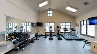 This fitness center is housed within a single-story space distinguished by a striking vaulted ceiling that creates an open, airy atmosphere. Skylights run along the roofline, supplementing the natural light that streams in through multiple large, grid-paned windows positioned along the exterior walls. The combination of overhead and ambient natural lighting ensures the space feels bright and energizing throughout the day.

The flooring consists of durable, speckled rubber or composite material well-suited for high-traffic exercise use. Along one wall, a full-length mirror extends the perceived depth of the room and aids exercisers in monitoring their form. A wall-mounted flat-screen television is positioned nearby for entertainment during workouts.

The cardio section is well-appointed, featuring treadmills, an elliptical trainer, a stationary upright bike, and a rowing machine. A dedicated free weights area lines one side of the space, offering a tiered dumbbell rack alongside medicine balls and battle ropes. The neutral, cream-toned walls maintain a clean, functional aesthetic throughout. Overall, the space is designed to support a comprehensive fitness routine in a comfortable, well-lit environment.