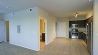 This single-floor residence offers a thoughtfully designed open-concept layout that seamlessly connects the main living areas. The space is finished with light-toned hardwood-style flooring that flows throughout the main areas, complemented by soft carpet in select zones, creating a warm and cohesive aesthetic.

The kitchen is a standout feature, equipped with stainless steel appliances including a range and built-in microwave, set against dark cabinetry with a tile backsplash. A ceiling fan with integrated lighting provides both comfort and illumination to the adjacent living space.

The open living and dining area benefits from a bright, airy atmosphere, thanks to neutral light-gray walls and multiple ceiling-mounted HVAC vents that ensure year-round comfort. The layout allows for flexible furniture arrangements, giving the space a versatile and functional character.

A bathroom is conveniently accessible from the main living area, featuring dark vanity cabinetry and a clean, functional design. A separate interior door provides access to additional private areas of the residence.

Overall, the space exudes a modern, minimalist ambiance with a neutral color palette, clean lines, and contemporary finishes that make it move-in ready and highly adaptable to a variety of interior styles.