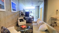 This single-floor space showcases a compelling industrial-chic aesthetic, defined by its exposed raw concrete walls and ceilings that lend a bold, urban character throughout. The open living area is generously proportioned, offering a comfortable and versatile layout. Polished concrete floors run throughout, reinforcing the loft-inspired design language and providing a sleek, low-maintenance foundation.

The living area serves as the centerpiece of the space, furnished with a plush cream sofa and coordinating armchair, centered around a glass-top coffee table resting on a patterned area rug. A wall-mounted flat-screen television is positioned on a black metal open shelving unit, which also serves as a display and storage solution. Natural light filters in through a bright window at the far end of the room, softening the industrial materials and creating a warm, inviting ambiance.

A ceiling fan is mounted overhead, adding both function and a subtle nod to classic loft living. A sliding door — finished in a contrasting warm yellow tone — adds a pop of color and a practical space-saving element. The overall atmosphere balances raw, utilitarian architecture with curated, comfortable furnishings, making it a distinctive and characterful space well-suited for modern urban living.