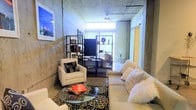 This single-floor space showcases a compelling industrial-chic aesthetic, defined by its exposed raw concrete walls and ceilings that lend a bold, urban character throughout. The open living area is generously proportioned, offering a comfortable and versatile layout. Polished concrete floors run throughout, reinforcing the loft-inspired design language and providing a sleek, low-maintenance foundation.

The living area serves as the centerpiece of the space, furnished with a plush cream sofa and coordinating armchair, centered around a glass-top coffee table resting on a patterned area rug. A wall-mounted flat-screen television is positioned on a black metal open shelving unit, which also serves as a display and storage solution. Natural light filters in through a bright window at the far end of the room, softening the industrial materials and creating a warm, inviting ambiance.

A ceiling fan is mounted overhead, adding both function and a subtle nod to classic loft living. A sliding door — finished in a contrasting warm yellow tone — adds a pop of color and a practical space-saving element. The overall atmosphere balances raw, utilitarian architecture with curated, comfortable furnishings, making it a distinctive and characterful space well-suited for modern urban living.