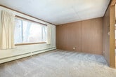 This single-floor, 2-bedroom, 1-bathroom unit offers a straightforward and functional layout across a compact footprint. The living room serves as the central gathering space, featuring wall-to-wall carpet, wood-paneled accent walls, a textured tile ceiling, and a large picture window that admits natural light and offers outdoor views of the surrounding neighborhood. Baseboard heating units run along the walls throughout the unit, providing consistent warmth.

The kitchen is equipped with warm-toned wood cabinetry offering ample storage, light-colored countertops, a coil-top electric range, and a full-sized refrigerator. Light wood-style flooring adds a clean, practical surface underfoot. An entry hallway with light flooring connects the main spaces and provides access to all rooms.

The primary bedroom includes carpeted flooring, a closet, a window with curtains, and baseboard heating. The second bedroom similarly features carpet, a window overlooking the exterior, and a baseboard heater. The bathroom is appointed with small-tile flooring, a single-sink vanity with cabinet storage, a toilet, and a tub-shower combination with a curtain. A skylight provides additional overhead light.