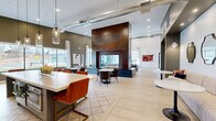 Located at 6 Crossgates Mall Road in Albany, NY, this thoughtfully designed amenity space spans a generous single floor, offering a sophisticated blend of modern industrial and contemporary design. The main lounge area is anchored by a dramatic floor-to-ceiling double-sided fireplace clad in rich Corten steel, serving as the visual centerpiece of the space. Light hardwood flooring flows throughout, complemented by soaring ceilings with exposed ductwork that add an urban industrial character. Large black-framed windows and glass doors flood the interior with natural light and provide views of an outdoor pool area. Multiple seating vignettes are thoughtfully arranged, including tufted leather sofas, upholstered chairs, and bistro-style dining sets. Built-in white shelving units flank a stylish wet bar with a beverage cooler, creating an ideal social hub. A dedicated game room features a billiards table and wall-mounted televisions, while a state-of-the-art golf simulator room is equipped with a projector, full-size screen, and putting surface. A quieter lounge nook with patterned carpeting and elegant drapery offers a more intimate retreat. The overall ambiance is polished, inviting, and well-appointed throughout.