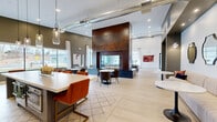 Located at 6 Crossgates Mall Road in Albany, NY, this thoughtfully designed amenity space spans a generous single floor, offering a sophisticated blend of modern industrial and contemporary design. The main lounge area is anchored by a dramatic floor-to-ceiling double-sided fireplace clad in rich Corten steel, serving as the visual centerpiece of the space. Light hardwood flooring flows throughout, complemented by soaring ceilings with exposed ductwork that add an urban industrial character. Large black-framed windows and glass doors flood the interior with natural light and provide views of an outdoor pool area. Multiple seating vignettes are thoughtfully arranged, including tufted leather sofas, upholstered chairs, and bistro-style dining sets. Built-in white shelving units flank a stylish wet bar with a beverage cooler, creating an ideal social hub. A dedicated game room features a billiards table and wall-mounted televisions, while a state-of-the-art golf simulator room is equipped with a projector, full-size screen, and putting surface. A quieter lounge nook with patterned carpeting and elegant drapery offers a more intimate retreat. The overall ambiance is polished, inviting, and well-appointed throughout.