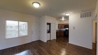 This single-floor unit presents an open-concept layout that seamlessly connects the living area to the kitchen, creating a cohesive and functional living environment. Dark hardwood-style flooring runs throughout, providing a warm and grounded aesthetic that contrasts with the crisp white walls and trim.

The living area is bright and straightforward, benefiting from a window that allows natural light to filter in. Flush-mount ceiling light fixtures illuminate the space consistently across both the main living zone and the kitchen area.

The kitchen is positioned toward the rear of the unit and is equipped with warm-toned wood cabinetry, dark countertops, and black appliances, including a range and overhead microwave. The layout provides ample functional workspace for everyday cooking needs.

A notable feature is the in-unit laundry closet, tucked neatly between the living area and kitchen. The closet houses a side-by-side washer and dryer behind paneled doors, offering added convenience. A secondary door near the laundry closet provides additional access or storage options. Overall, the space is clean, practical, and move-in ready.