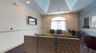 This single-floor space functions as a comfortable lounge or common gathering area, thoughtfully designed to accommodate relaxed seating and entertainment. A defining architectural feature is the tray ceiling, which adds visual depth and dimension to the room while recessed lighting provides a warm, even illumination throughout. An arched window fitted with sheer white curtains serves as a natural focal point on the far wall, allowing diffused natural light to brighten the interior.

The seating arrangement includes a row of patterned upholstered chairs in the foreground, featuring a geometric design in earth tones of brown and teal, complemented by a beige sofa positioned against the back wall and accented with decorative throw pillows in dark and teal hues. A wall-mounted flat-screen television is positioned prominently on one wall, reinforcing the space's function as a media and social lounge. A classic accent chair in dark wood is visible near the window, adding a subtle design contrast. The neutral wall tones, carpeted flooring, and layered lighting contribute to an overall ambiance that is both functional and inviting.