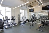 This comprehensive fitness center is thoughtfully designed to accommodate a wide range of workout styles and training needs. The space is divided into distinct zones, each tailored for specific activities. The main cardio and strength training area features durable rubber flooring, floor-to-ceiling windows that flood the space with natural light, and views of the surrounding exterior. This zone is outfitted with treadmills, ellipticals, cable machines, weight benches, and a full dumbbell rack, all complemented by wall-mounted mirrors and flat-screen TVs. An oversized ceiling fan adds comfort to the high-ceilinged space, which showcases exposed ductwork and industrial-style linear lighting for a modern aesthetic. Adjacent to the main floor is a dedicated functional training and studio area finished with rich hardwood flooring. This versatile zone includes TRX suspension trainers mounted to a wall-mounted pull-up rig, a BOSU ball, stacked plyo boxes, and a color-coded dumbbell set. A full-length mirror wall, spin bikes, rowing machines, stability balls, and a wall-mounted TV make this area ideal for group fitness or independent training. Built-in open shelving provides convenient storage throughout the studio space.
