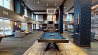 This expansive amenity lounge is designed with a contemporary aesthetic that blends warmth and sophistication throughout its open-plan layout. Soaring double-height ceilings with recessed and linear lighting create a dramatic sense of volume, while bold textured columns clad in dark speckled stone anchor the space visually and add architectural character.

At the heart of the lounge, a full-size billiards table with a teal felt surface sits atop a herringbone-patterned hardwood inlay, complemented by a shuffleboard table nearby, making this a standout destination for recreation. A striking floor-to-ceiling stacked stone fireplace with a linear gas insert serves as the focal point of the main seating area, flanked by leather sofas and a center coffee table, with a wall-mounted television above.

Multiple distinct seating zones are thoughtfully arranged throughout the space, including a casual lounge area with large floor-to-ceiling windows dressed in gold drape panels that invite natural light and outdoor views. Additional seating nooks with leather armchairs and game tables provide intimate gathering spots. A staircase with wood treads and black metal railings leads to an upper-level overlook, adding vertical dimension. Patterned carpet throughout ties the various zones together, while warm wood tones and dark accent walls contribute to the overall inviting and upscale ambiance.