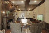 This single-floor space showcases a refined blend of traditional craftsmanship and lodge-inspired design. The living area is defined by striking dark wood coffered ceilings that add architectural depth and visual drama overhead. Rich hardwood flooring runs throughout, complementing extensive dark wood wainscoting that lines the walls, which are finished in a textured grasscloth-style wall covering above.

A prominent fireplace with an ornate dark wood surround serves as a commanding focal point, topped by a mounted moose head that reinforces the space's sophisticated rustic character. Framed artwork and decorative wall-mounted pieces add curated visual interest throughout. The sitting area is thoughtfully arranged with upholstered accent chairs, a tufted leather ottoman, and a flat-screen television set within a handsome dark wood media console.

Large French doors with dark wood frames and transom windows above create a dramatic transition between rooms, flooding the interior with natural light while maintaining a sense of defined spaces. Beyond the French doors, a brighter, lighter-toned area with tile flooring is visible, offering a pleasing contrast to the warm, wood-rich primary living space. Recessed lighting throughout ensures the rooms remain well-illuminated and welcoming.