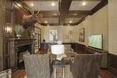 This single-floor space showcases a refined blend of traditional craftsmanship and lodge-inspired design. The living area is defined by striking dark wood coffered ceilings that add architectural depth and visual drama overhead. Rich hardwood flooring runs throughout, complementing extensive dark wood wainscoting that lines the walls, which are finished in a textured grasscloth-style wall covering above.

A prominent fireplace with an ornate dark wood surround serves as a commanding focal point, topped by a mounted moose head that reinforces the space's sophisticated rustic character. Framed artwork and decorative wall-mounted pieces add curated visual interest throughout. The sitting area is thoughtfully arranged with upholstered accent chairs, a tufted leather ottoman, and a flat-screen television set within a handsome dark wood media console.

Large French doors with dark wood frames and transom windows above create a dramatic transition between rooms, flooding the interior with natural light while maintaining a sense of defined spaces. Beyond the French doors, a brighter, lighter-toned area with tile flooring is visible, offering a pleasing contrast to the warm, wood-rich primary living space. Recessed lighting throughout ensures the rooms remain well-illuminated and welcoming.