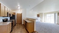 This single-floor unit offers a well-designed open-concept layout that seamlessly connects the kitchen and living areas. The kitchen is outfitted with warm honey-toned wood cabinetry, a peninsula with a built-in sink and granite-style countertops, a stainless steel refrigerator, and a dishwasher. Hardwood-style flooring runs throughout the kitchen and hallway, providing a clean and durable surface. The peninsula serves as a natural divider between the kitchen and the adjoining living space, which transitions to plush wall-to-wall carpeting for added comfort underfoot. The living area is generously proportioned and benefits from natural light streaming through a large window, creating a bright and welcoming atmosphere. Neutral wall tones throughout contribute to a calm, versatile interior that suits a variety of furnishing styles. Interior doors feature classic six-panel wood designs with warm wood trim, adding a traditional detail to the space. A baseboard heating system is visible along the walls of the living area, ensuring year-round comfort. The layout provides a functional flow, with clearly defined zones for cooking, dining, and relaxing, making this a practical and inviting single-level space.