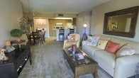 This single-floor residence features an open-concept design that seamlessly connects the main living areas, creating a cohesive and functional flow throughout the space. The living room serves as the central gathering area, furnished with a neutral-toned sofa and accent chair accented with floral throw pillows, anchored by a wooden glass-top coffee table. Soft carpeting underfoot adds warmth to the space, while a floor lamp provides ambient lighting. The living area opens directly into a defined dining space, which comfortably accommodates a full dining table and chair set, ideal for everyday meals or entertaining. The kitchen is positioned at the rear of the open layout and features warm wood-toned cabinetry, a white refrigerator, and a raised breakfast bar with bar seating — offering both additional prep space and casual dining options. The kitchen benefits from its own overhead lighting, keeping the cooking area well-illuminated. Throughout the space, the open sightlines between rooms enhance the sense of spaciousness, making the layout feel connected and inviting. A ceiling fan is also visible, contributing to year-round comfort in the main living area.