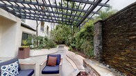 This thoughtfully designed outdoor space offers a comprehensive resort-style experience within a landscaped courtyard setting. A striking black steel pergola anchors a comfortable lounge area furnished with wicker seating, navy cushions, and a teak coffee table, positioned alongside curved concrete bench walls that wind through densely planted garden beds filled with bamboo, palms, and flowering shrubs. The freeform swimming pool serves as a central feature, its turquoise water surrounded by a broad concrete deck outfitted with lounge chairs, bistro sets, and striped umbrellas. Adjacent to the pool, a fully equipped outdoor kitchen features a stainless steel built-in grill, counter space, and storage, accompanied by a dining area with teak table and chairs. A dramatic stacked-slate water feature wall adds a sculptural focal point, with natural stone coping and river rock detailing at its base. Large cylindrical planters throughout the open courtyard plaza provide visual interest and greenery. French doors along the building facade connect the outdoor space to interior amenities, reinforcing the seamless indoor-outdoor flow that defines the overall ambiance.