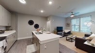 This single-floor residence is designed with a contemporary aesthetic, featuring neutral gray walls, wood-look plank flooring, and clean-lined finishes throughout the main living areas. The open-concept kitchen is a central highlight, anchored by a white quartz island with a double-basin sink and bar seating. Two-tone cabinetry in a warm wood-grain finish provides ample storage, while stainless steel appliances—including a refrigerator, range, and microwave—complete the functional workspace. Recessed lighting illuminates the kitchen and entry corridor effectively.

The kitchen flows seamlessly into the living area, which benefits from natural light through window blinds and is outfitted with a ceiling fan. A dedicated sitting nook near the window offers a comfortable secondary space for relaxing or working.

The bedroom features plush carpeting, a ceiling fan, and a window that allows natural light to fill the room. The bathroom is notably spacious, presenting a long, corridor-style layout with wood-look flooring, a single vanity with quartz countertop, and access to a walk-in closet with open rod storage. A curtained shower/tub combination completes the bathroom. Throughout the unit, consistent finishes and neutral tones create a cohesive, modern living environment.