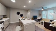 This single-floor residence is designed with a contemporary aesthetic, featuring neutral gray walls, wood-look plank flooring, and clean-lined finishes throughout the main living areas. The open-concept kitchen is a central highlight, anchored by a white quartz island with a double-basin sink and bar seating. Two-tone cabinetry in a warm wood-grain finish provides ample storage, while stainless steel appliances—including a refrigerator, range, and microwave—complete the functional workspace. Recessed lighting illuminates the kitchen and entry corridor effectively.

The kitchen flows seamlessly into the living area, which benefits from natural light through window blinds and is outfitted with a ceiling fan. A dedicated sitting nook near the window offers a comfortable secondary space for relaxing or working.

The bedroom features plush carpeting, a ceiling fan, and a window that allows natural light to fill the room. The bathroom is notably spacious, presenting a long, corridor-style layout with wood-look flooring, a single vanity with quartz countertop, and access to a walk-in closet with open rod storage. A curtained shower/tub combination completes the bathroom. Throughout the unit, consistent finishes and neutral tones create a cohesive, modern living environment.