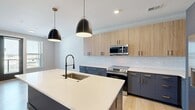 Located at 1805 Boulevard W in Richmond, Virginia, this thoughtfully designed one-bedroom, one-bathroom residence offers a modern aesthetic with high-quality finishes across its single floor. The open-concept kitchen serves as the centerpiece of the home, showcasing a striking two-tone cabinetry scheme — slate-blue lower cabinets with brushed gold hardware paired with light wood upper cabinets. A spacious quartz-topped island with an integrated sink and sleek matte black pendant lighting anchors the space, complemented by a white subway tile backsplash and a full suite of stainless steel appliances, including a gas range and French door refrigerator. Light hardwood flooring flows throughout the main living areas, enhancing the bright, airy atmosphere. The dining area benefits from natural light streaming through large windows with views of the surrounding neighborhood. The bedroom is a calm retreat, featuring plush carpeting, a ceiling fan, and generous windows that invite abundant daylight. The bathroom carries the home's cohesive design language with navy cabinetry, gold hardware, white quartz countertops, and a full tub-shower combo with matte black fixtures and a bold gray shaker-style door.