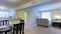 This single-floor residence offers a well-designed open-concept layout that efficiently connects the main living areas. The kitchen is positioned at one end, featuring light-toned cabinetry, granite or stone-look countertops, and built-in appliances including a microwave. A breakfast bar serves as a natural divider between the kitchen and the adjacent dining area, which comfortably accommodates a dark-wood dining set. The open floor plan flows seamlessly into the spacious living room, where plush wall-to-wall carpeting adds warmth underfoot. The living area is bathed in natural light from a large window that overlooks the outdoors, creating a bright and inviting atmosphere. Neutral white and soft gray walls throughout the space lend a clean, modern aesthetic that complements a variety of décor styles. Structural beams along the ceiling add a subtle architectural detail, while multiple HVAC vents ensure consistent climate control. A doorway visible from the main living area provides access to additional rooms, suggesting a thoughtful and functional layout. Overall, the space conveys a comfortable, move-in-ready environment with a cohesive and understated design sensibility.