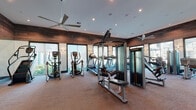 This contemporary fitness center is thoughtfully designed to support a full range of workout routines across multiple connected zones. The space is unified by a warm, sophisticated aesthetic, highlighted by rich wood-paneled accent walls, recessed lighting, and large ceiling fans that maintain a comfortable environment throughout.

The primary cardio area is lined with a fleet of treadmills and elliptical machines, all equipped with digital screens and positioned in front of floor-to-ceiling windows that invite abundant natural light and offer views of the surrounding outdoor amenity areas. Mirrored walls further amplify the sense of openness in this zone.

Adjacent to the cardio area is a dedicated strength training section housing a variety of cable and resistance machines, including a functional trainer and seated press equipment, complemented by Spirit rowing machines for a full-body workout.

A separate functional training studio offers hardwood flooring, stability balls in multiple sizes, medicine ball racks, resistance bands, and a foam roller station — making it ideal for group exercise, stretching, or dynamic movement work. Large glass panel windows with privacy shades connect this studio to the main floor while preserving flexibility of use.

Throughout the facility, the design balances practicality with a polished, upscale ambiance, making it a well-rounded destination for fitness of all kinds.