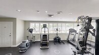 This fitness center in Carrboro, NC offers a dedicated exercise room designed to support a well-rounded workout routine. The space features a dark, durable flooring surface that is well-suited for high-traffic use, complemented by light sage-green walls and crisp white trim that create a clean, focused atmosphere. Recessed lighting throughout ensures consistent illumination across the entire floor area.

The exercise room is generously outfitted with a wide variety of equipment, including multiple treadmills, an elliptical trainer, a stair climber, stationary and spin bikes, and a multi-station strength training machine. A wall-mounted flat-screen television adds an entertainment option for users during their workouts.

Large windows fitted with blinds line one wall, allowing ample natural light to fill the space while maintaining privacy. A glass-paneled door provides direct access to an outdoor area, offering views of the surrounding neighborhood. Additional doors lead to separate utility or storage rooms, and a wall-mounted hand sanitizer station supports cleanliness and hygiene. The overall layout is practical and spacious, providing enough room to move freely between equipment while maintaining a comfortable, gym-like environment.