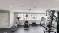 This fitness center in Carrboro, NC offers a dedicated exercise room designed to support a well-rounded workout routine. The space features a dark, durable flooring surface that is well-suited for high-traffic use, complemented by light sage-green walls and crisp white trim that create a clean, focused atmosphere. Recessed lighting throughout ensures consistent illumination across the entire floor area.

The exercise room is generously outfitted with a wide variety of equipment, including multiple treadmills, an elliptical trainer, a stair climber, stationary and spin bikes, and a multi-station strength training machine. A wall-mounted flat-screen television adds an entertainment option for users during their workouts.

Large windows fitted with blinds line one wall, allowing ample natural light to fill the space while maintaining privacy. A glass-paneled door provides direct access to an outdoor area, offering views of the surrounding neighborhood. Additional doors lead to separate utility or storage rooms, and a wall-mounted hand sanitizer station supports cleanliness and hygiene. The overall layout is practical and spacious, providing enough room to move freely between equipment while maintaining a comfortable, gym-like environment.