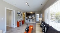 This single-floor residence showcases a contemporary open-concept design that seamlessly connects the living and kitchen areas. The living space is anchored by light hardwood-style flooring and neutral gray walls, providing a versatile backdrop for bold décor accents, including vibrant orange furnishings and striking wall art. Track lighting mounted on the ceiling illuminates the space efficiently, highlighting its clean, modern aesthetic.

The kitchen is fitted with dark cabinetry contrasting against lighter countertops, stainless steel appliances, and a breakfast bar equipped with sleek black pedestal stools — ideal for casual dining or entertaining. A door adjacent to the kitchen area leads to an outdoor space, allowing natural light to filter in.

The living area features a comfortable seating arrangement and is accessorized with decorative pillows and table lamps, creating an inviting atmosphere. Large windows with blinds allow natural light throughout the unit.

From an elevated position, the unit offers expansive views of a city skyline, with high-rise buildings and a wide urban landscape stretching beneath a clear blue sky, providing a compelling urban backdrop to daily living.