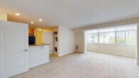 This single-floor residence offers a well-organized, open-concept layout that efficiently connects the kitchen, dining, and living areas into one cohesive space. The kitchen is equipped with warm honey-toned wood cabinetry, dark granite-style laminate countertops, a stainless steel sink, and black appliances including an electric range, over-the-range microwave, and refrigerator. A breakfast bar peninsula separates the kitchen from the main living area, providing additional counter space and a casual dining option. The living area is generously proportioned, finished with plush neutral carpeting, and bathed in natural light through a wide bank of windows. Recessed lighting throughout the common areas adds to the bright, clean ambiance. A dedicated in-unit laundry closet houses a stacked washer and dryer, adding everyday convenience. The bathroom features tile flooring, a soaking bathtub with a shower curtain rod, brushed nickel hardware accents, and clean white walls. Additional storage is accessible via a closet near the entry. The overall design is neutral and move-in ready, with a light color palette and clean lines that provide a versatile backdrop for any furnishing style.