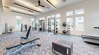 Located at 2307 Ridgemont Ave in Charlotte, NC, this fitness center offers a generously sized, single-floor exercise space designed to support a full range of training activities. The room features white shiplap walls, a coffered ceiling with recessed lighting and ceiling fans, and large multi-pane windows that flood the space with natural light while offering views of the surrounding neighborhood.

The strength training area is thoroughly outfitted with a multi-functional cable machine, a power rack with a full set of weight plates, a dumbbell rack, medicine ball storage, and adjustable benches. A plyo box and TRX-style suspension equipment further enhance functional fitness options. The cardio zone along the windowed wall includes a row of treadmills and upright cardio machines, each equipped with individual display screens. A wall-mounted television provides additional entertainment.

Stylish blue-toned patterned carpet tiles cover the floor throughout, adding visual interest while providing a durable surface for varied workouts. Near the entrance, a leg press machine and a small welcome station with decorative elements greet users. Additional ancillary rooms are accessible off the main floor, potentially serving as changing or utility spaces. The overall ambiance is bright, modern, and purpose-built for an active lifestyle.