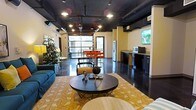 This single-floor space showcases a striking industrial-chic aesthetic, seamlessly blending raw architectural elements with contemporary furnishings. The exposed ceiling reveals ductwork and conduit painted in dark tones, complemented by track lighting and pendant fixtures that illuminate the open-plan interior. Polished concrete floors reflect light throughout, enhancing the spacious feel of the layout.

The living area anchors the space with a teal sofa, a round dark-wood coffee table atop a gold-and-cream patterned area rug, and carefully curated décor accents. A large garage-style glass door/window wall at the far end floods the space with natural light and provides views to an outdoor area, where vibrant red seating is visible.

The kitchen and service area along one wall features dark cabinetry with granite countertops, a stainless steel refrigerator, microwave, and a wall-mounted flat-screen television above, creating a functional yet stylish zone. Lush indoor plants near the window wall add a refreshing natural contrast to the industrial elements. Overall, the space exudes a modern, versatile ambiance suited for both work and leisure.