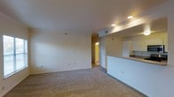 This single-floor residential unit offers a clean, functional layout designed for comfortable everyday living. The open-concept living area is spacious and filled with a sense of openness, featuring neutral wall tones and plush wall-to-wall carpeting that adds warmth and comfort underfoot. Recessed lighting throughout the main living space provides consistent, even illumination, enhancing the bright and airy atmosphere of the interior.

Adjacent to the living area, the kitchen is defined by a raised breakfast bar counter that serves as both a practical workspace and a casual dining or gathering spot. White cabinetry lines the kitchen, offering ample storage while maintaining a clean, cohesive aesthetic. The kitchen and living areas flow naturally into one another, making the space well-suited for entertaining or daily routines.

A hallway visible from the main living area leads to the remaining rooms of the unit, which are warmly lit and accessible. HVAC vents are integrated into the ceiling, ensuring year-round climate control. The overall design is understated and versatile, offering a neutral canvas that can be easily personalized to suit a variety of interior styles. The single-floor layout ensures seamless accessibility and ease of movement throughout the entire space.