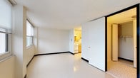This single-floor unit offers a straightforward and functional layout designed for comfortable everyday living. The main living area is open and well-lit, featuring large windows fitted with horizontal blinds that allow ample natural light to fill the space while offering views of surrounding greenery. The polished vinyl tile flooring runs throughout, providing durability and easy maintenance. Bold black baseboards provide a clean contrast against the white walls, adding a subtle architectural detail throughout.

The compact kitchen is efficiently designed with light wood cabinetry offering generous storage, a stainless steel sink, a white electric range with coil burners, and a full-size refrigerator. An overhead range hood and a textured ceiling complete the kitchen's practical setup.

The bathroom is neatly appointed with white tile surrounding the full bathtub and shower, a mosaic tile floor, a wall-mounted sink, a mirrored medicine cabinet, and a standard toilet. The clean, white palette creates a bright and hygienic feel.

The main living area also features multiple doors leading to closet storage and the bathroom, providing organizational convenience. A wall-mounted HVAC unit ensures climate control throughout the space. Overall, the unit presents a clean, no-frills environment well-suited for functional, day-to-day use.