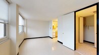 This single-floor unit offers a straightforward and functional layout designed for comfortable everyday living. The main living area is open and well-lit, featuring large windows fitted with horizontal blinds that allow ample natural light to fill the space while offering views of surrounding greenery. The polished vinyl tile flooring runs throughout, providing durability and easy maintenance. Bold black baseboards provide a clean contrast against the white walls, adding a subtle architectural detail throughout.

The compact kitchen is efficiently designed with light wood cabinetry offering generous storage, a stainless steel sink, a white electric range with coil burners, and a full-size refrigerator. An overhead range hood and a textured ceiling complete the kitchen's practical setup.

The bathroom is neatly appointed with white tile surrounding the full bathtub and shower, a mosaic tile floor, a wall-mounted sink, a mirrored medicine cabinet, and a standard toilet. The clean, white palette creates a bright and hygienic feel.

The main living area also features multiple doors leading to closet storage and the bathroom, providing organizational convenience. A wall-mounted HVAC unit ensures climate control throughout the space. Overall, the unit presents a clean, no-frills environment well-suited for functional, day-to-day use.