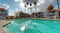Located at 3202 W Bell Rd in Phoenix, Arizona, this single-story community showcases a distinctly Southwestern architectural style, characterized by stucco exteriors in cool grey tones, terracotta-tiled rooflines, and clean geometric facades. The buildings are arranged around a central outdoor amenity area that serves as the heart of the community.

The centerpiece of the outdoor space is a generously sized swimming pool with vivid turquoise water, framed by a wide concrete pool deck that provides ample room for lounging. Rows of chaise lounge chairs line the perimeter, offering residents dedicated spaces to relax and enjoy the Arizona sunshine. A covered pergola structure along one side of the deck provides shaded seating, ideal for escaping the desert heat while remaining close to the pool.

Towering palm trees rise dramatically above the roofline, reinforcing the desert-resort ambiance of the property. The surrounding buildings feature gated access points, iron fencing, and stairway entries that connect directly to the pool area. A spa or hot tub is also present near the pool's edge, adding another layer of outdoor leisure. The overall atmosphere is open, sun-drenched, and resort-like, capturing the relaxed outdoor lifestyle characteristic of Phoenix living.