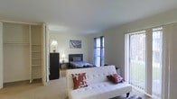 This single-floor space is designed as an open-concept layout that seamlessly integrates the living and sleeping areas into one cohesive environment. The room is finished with neutral-toned walls and carpeted flooring, creating a clean and understated aesthetic throughout.

The sleeping area is positioned toward the back of the space and features a twin bed dressed in a patterned comforter, accompanied by a compact dresser and a bedside table with a lamp, providing functional storage and ambient lighting. A framed piece of wall art adds a decorative touch above the bed. A window dressed with blue curtains allows natural light to filter into this area.

The living area is anchored by a white tufted futon accented with decorative throw pillows, offering flexible seating and additional sleeping capacity. Large sliding glass doors with vertical blinds flood the space with abundant natural light while providing direct access to an outdoor area.

To one side, open shelving and additional storage solutions are partially visible, contributing to the overall functionality of the layout. The space is compact yet well-organized, making efficient use of every area while maintaining a comfortable and livable atmosphere.