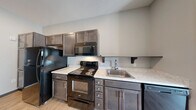 Located at 3080 E Cherry St in Springfield, Missouri, this one-bedroom, one-bathroom unit offers a smartly designed single-level layout that maximizes every square foot. The open-concept kitchen flows directly into the living area, creating a connected and functional living space. The kitchen is well-appointed with dark espresso cabinetry extending to the ceiling for generous storage, light-toned countertops, and a full suite of black Whirlpool appliances including a refrigerator, electric range, over-the-range microwave, and dishwasher. Light wood-style flooring runs throughout the main living areas, lending a clean and contemporary aesthetic. The living room receives natural light through a sliding glass door that opens to a private balcony, offering an outdoor retreat. The bedroom features neutral carpet, a window with blinds, and direct access to a dedicated closet. The bathroom is finished with wood-style flooring and includes a full tub-shower combination with a sliding glass enclosure. Additional utility and laundry rooms provide practical in-unit convenience. Recessed lighting and a neutral color palette throughout give the space a modern, move-in-ready feel.