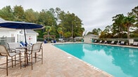 Located at 1220 Meredith Dr in Columbia, South Carolina, this outdoor amenity space centers around a generously sized swimming pool with crystal-clear turquoise water. The pool ranges from a shallow end to a depth of four feet, making it accessible for a variety of swimmers. Surrounding the pool is a wide, brick-paved pool deck that provides ample room for lounging, socializing, and outdoor recreation.

A well-appointed pool house anchors one side of the space, featuring an arched window, a pitched roof, and a classic exterior design that complements the overall aesthetic of the facility. Along the perimeter of the deck, rows of lounge chairs and chaise lounges offer dedicated sunbathing spots.

A pergola-covered dining area provides a shaded retreat, furnished with tables and chairs ideal for outdoor gatherings. Adjacent to this is a cozy fire pit lounge area with Adirondack chairs set on artificial turf, offering a relaxed setting for evening socializing.

The entire pool area is enclosed by a black metal fence and framed by lush, mature landscaping that includes tall pine trees, palm trees, and manicured evergreen shrubs, creating a private and serene atmosphere. Decorative blue planters add vibrant accent touches throughout the space, enhancing the resort-like character of this well-designed outdoor amenity.