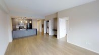 This single-floor residence presents an open-concept layout that seamlessly connects the living area to the kitchen, creating a spacious and functional environment. Light wood-tone laminate flooring extends throughout, lending warmth and continuity to the space.

The kitchen is positioned to the left and features dark stainless steel appliances, including a refrigerator, complemented by a sleek island or peninsula with a dark countertop and modern grey cabinetry. Pendant lighting hangs above, adding a contemporary touch to the cooking area.

The open living space is framed by neutral beige walls and a white ceiling, creating a bright and airy atmosphere. Recessed lighting supplements the overall illumination throughout the unit.

Toward the rear, a dedicated in-unit laundry area is tucked neatly between white cabinetry, offering convenient access without sacrificing style. Adjacent hallways lead to additional rooms, suggesting private spaces such as bedrooms or a bathroom. White interior doors and trim provide clean architectural contrast against the warm-toned walls, contributing to the space's modern and well-organized character.