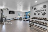 Located at 9651 Graham Street in Cypress, CA, this fitness center at Cypress Park is a well-appointed, single-room exercise facility designed to support a comprehensive workout routine. The space features durable rubber flooring throughout, recessed lighting, and a textured white ceiling that keeps the environment bright and functional. A bold teal accent stripe runs along the base of the walls, complemented by motivational wall graphics and Cypress Park branding that add personality to the room.

The facility is equipped with a variety of cardio machines, including a treadmill, elliptical, and stationary bike, as well as a multi-station cable strength machine and a rowing machine. A rack of dumbbells in graduated weights provides ample options for free weight training, and a workout bench supports a range of exercises. Wall-mounted resistance bands and yoga mats offer additional versatility.

Multiple windows along the walls allow natural light to filter in and provide views of the surrounding outdoor area. A wall-mounted flat-screen television offers entertainment during workouts. Full-length mirrors on one wall visually expand the space and assist with form. A water fountain with a bottle-fill station is conveniently positioned near the entrance. The room also displays posted fitness center rules, ensuring a safe and organized environment for all users.