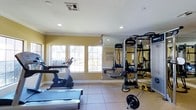 This single-floor fitness center is thoughtfully designed to support a well-rounded workout experience. The space features a clean, open layout finished with neutral-toned walls and light tile flooring, creating a bright and energizing atmosphere. Recessed lighting complements the natural light that floods in through a row of large, multi-pane windows along one wall, keeping the environment airy and inviting.

The cardio area is equipped with a treadmill, stationary bike, and a rowing machine, providing a solid range of options for aerobic exercise. On the strength training side, a robust multi-station cable and pulley system by Life Fitness anchors the room, accompanied by free weights including a barbell with weight plates and an assortment of medicine balls. Full-length mirrors line one wall, both enhancing the sense of space and allowing users to monitor their form during workouts.

Suspension training straps are also available, offering additional functional fitness options. The ceiling features a standard HVAC vent and smoke detector, ensuring comfort and safety throughout. The overall layout is practical and user-friendly, making efficient use of the available floor space while keeping equipment accessible and well-organized. This fitness center provides a comprehensive and comfortable environment for residents or users to meet their health and wellness goals.