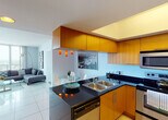 Located along the vibrant Biscayne Boulevard corridor in Miami, this contemporary high-rise residence offers a well-designed single-floor layout that seamlessly blends modern aesthetics with functional living. Floor-to-ceiling sliding glass doors and expansive windows flood the interior with natural light while framing sweeping panoramic views of the Miami skyline and surrounding cityscape.

The open-concept living and dining area is anchored by large-format porcelain tile flooring and features a generous sectional seating arrangement oriented toward the breathtaking views. A round glass dining table with modern white chairs sits adjacent to the kitchen, illuminated by striking orange pendant lights that add a bold design accent.

The kitchen is outfitted with warm honey-toned wood cabinetry, dark granite countertops, and stainless steel appliances including a built-in oven, microwave, refrigerator, and dishwasher — providing a complete and stylish culinary space.

The bedroom is a serene retreat with plush carpeting, soft blue-grey walls, and direct access to the balcony through floor-to-ceiling glass doors, continuing the theme of expansive city views. A full bathroom features tile flooring, a combined tub and shower with tile surround, and a warm-toned vanity countertop with an undermount sink. Throughout, the residence maintains a clean, contemporary palette that reflects Miami's cosmopolitan character.
