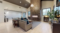 This single-story community clubhouse is designed with a contemporary aesthetic that balances comfort and functionality throughout its open, well-appointed layout. The centerpiece of the space is a dramatic lounge area distinguished by soaring vaulted ceilings, light tile flooring, and a striking floor-to-ceiling herringbone-patterned accent wall framing a linear gas fireplace. A chandelier adds an elegant touch overhead, while plush gray sofas and teal accent pillows create a welcoming seating arrangement. A mounted flat-screen television on a decorative media console provides additional comfort to the lounge. Adjacent to the lounge, a casual dining and gathering area offers seating at dark bistro-style tables beneath recessed lighting. A dedicated conference room features a large central table, ergonomic seating, a wall-mounted display screen, and expansive windows that invite abundant natural light and views of the surrounding community. A separate seating nook with a tufted leather sofa and framed wall art provides a quieter retreat. The space also includes a leasing office area and a sleek kitchenette outfitted with gray cabinetry, quartz countertops, open stainless steel shelving, a utility sink, and a beverage refrigerator — ideal for hosting residents and guests alike.
