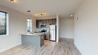 This single-floor unit offers a clean, contemporary design with an open-concept layout that seamlessly connects the kitchen and living area. The kitchen is a standout feature, equipped with stainless steel appliances including a refrigerator, electric range, and over-the-range microwave. Dark wood cabinetry contrasts elegantly with light granite countertops, while a mosaic tile backsplash adds a refined touch. A peninsula with an undermount sink and pendant lighting above serves as both a functional workspace and a natural divider between the kitchen and the open living space. Light wood-style flooring runs throughout the main living areas, lending warmth and continuity to the space. Large windows with horizontal blinds invite natural light into the living area, which also features access to an outdoor balcony. Track lighting throughout adds a modern, versatile illumination option. The two bedrooms are finished with soft, neutral carpet and each includes a closet and a window for natural light. The full bathroom is clean and well-appointed, featuring a tub and shower combination enclosed by sliding glass doors, a wall-mounted vanity with a white basin, a framed mirror, and neutral tile flooring, creating a bright and functional space overall.